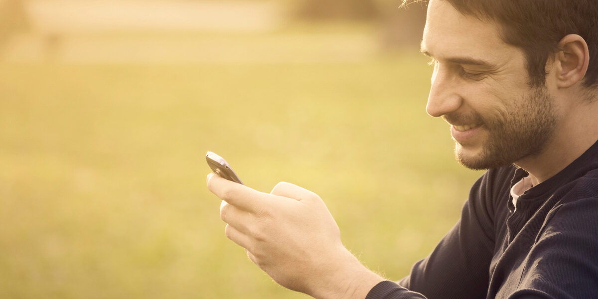 Ein junger Mann sitzt in der Natur und freut sich, am Handy eine Immobilie zum Kauf gefunden zu haben | Immobilienkauf