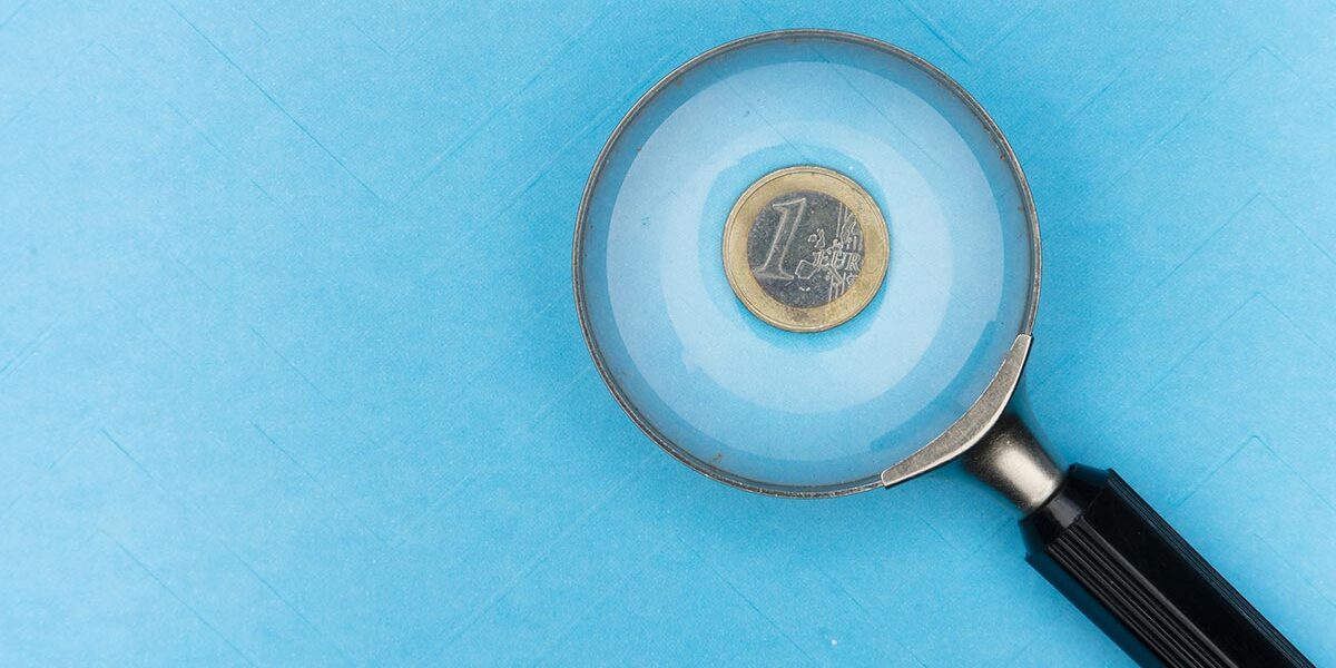 Auf einer blauen Unterlage, unter einer Lupe liegt eine 1-Euro-Münze, was symbolisiert, dass man bei der Immobilienfinanzierung ganz genau aufs Geld schauen muss | Immobilienfinanzierung